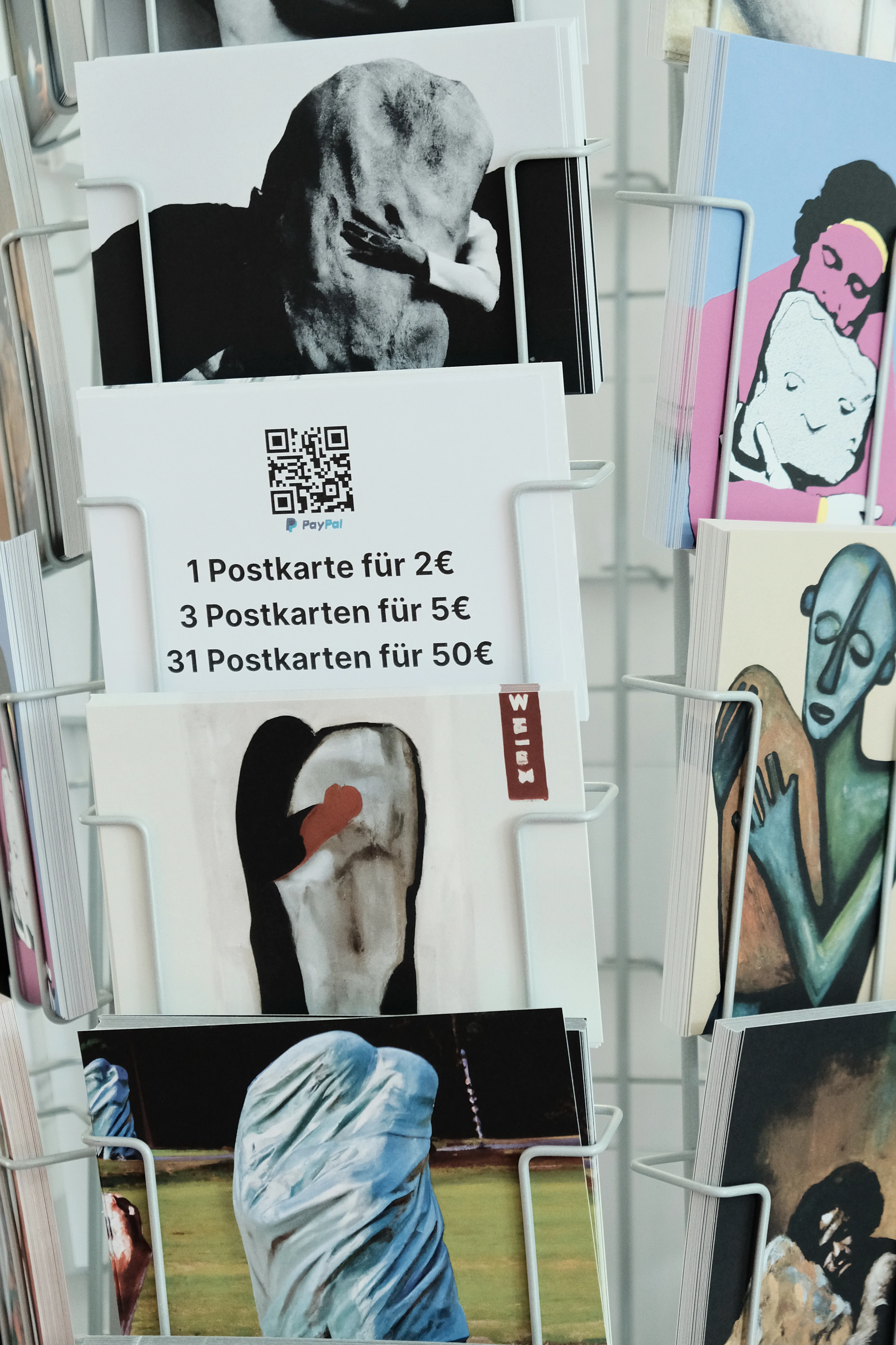 Close-up of a postcard stand displaying various artworks, including surreal and abstract depictions of figures embracing stones. A sign in the stand shows a QR-code labeled 'PayPal' and lists prices: 1 postcard for 2€, 3 for 5€, and 31 for 50€.