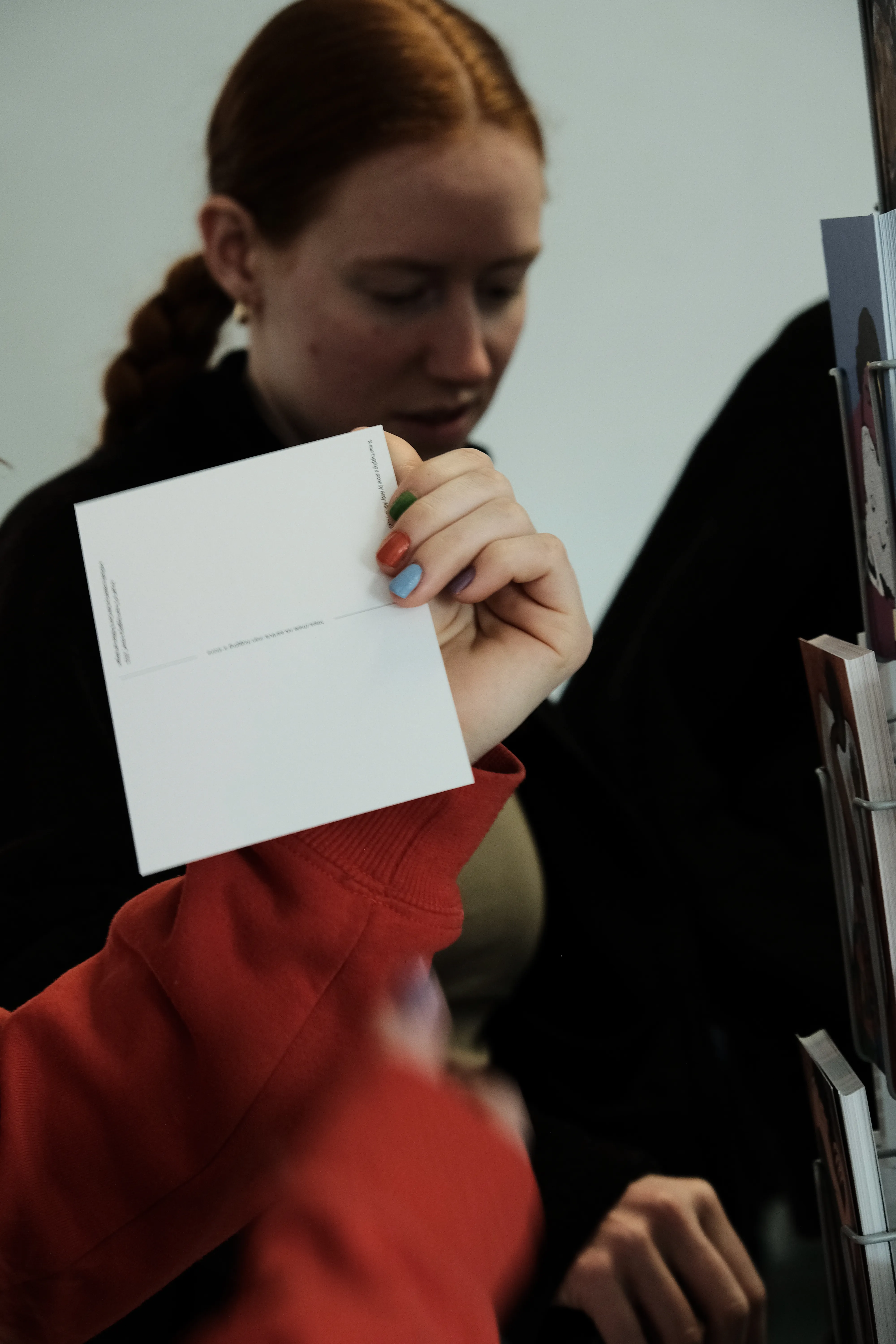 Closeup of a person's hand examining the back side of a postcard. The Postcard stand is visible and cut-off to the right. Another person is visible in the background reaching for the stand.