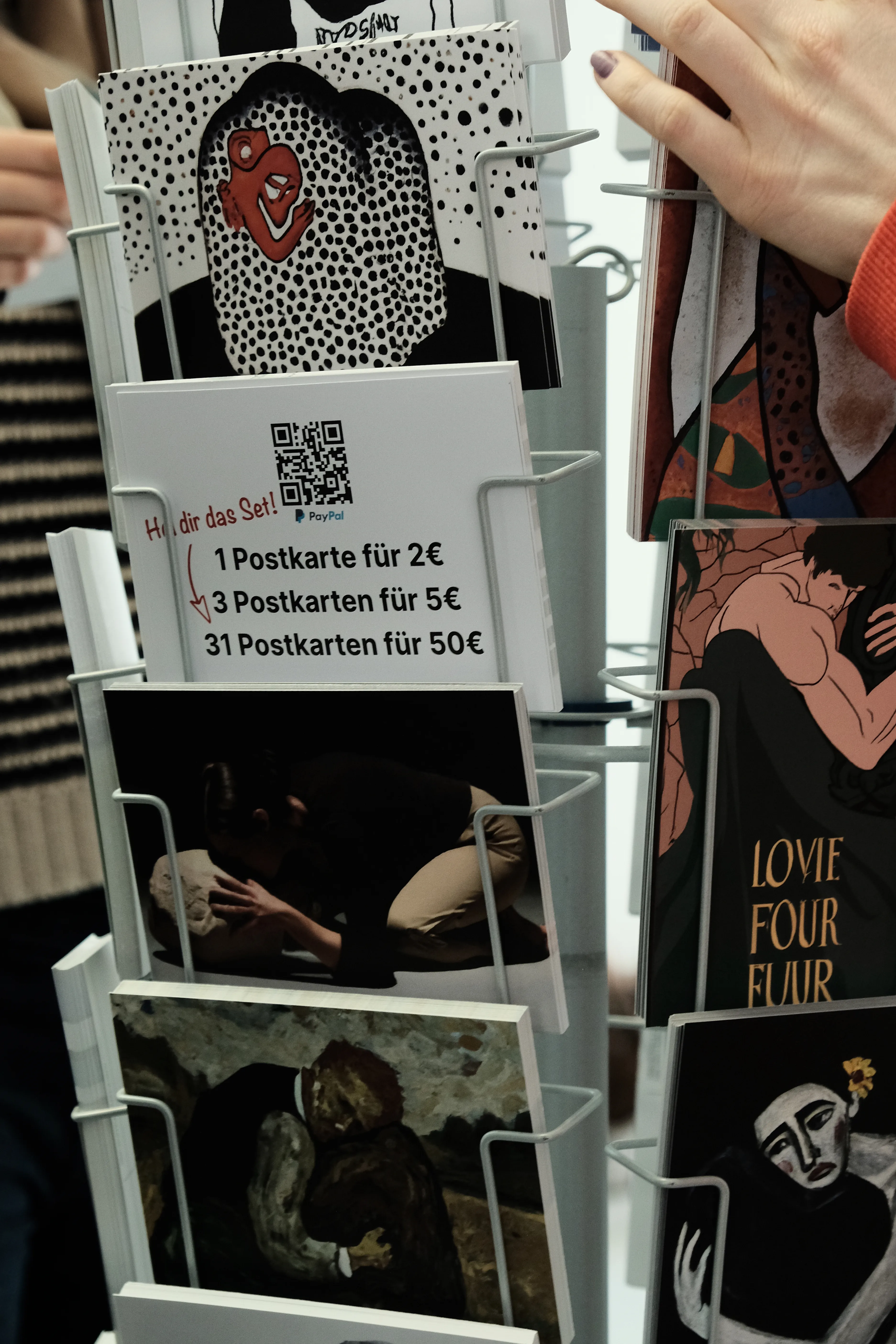 Close-up of a postcard stand displaying various artworks, including surreal and abstract depictions of figures embracing stones. A thin cardboard box the size of a postcard with an opening at the top is placed in the stand and shows an illustration of three coins and lists prices: 1 postcard for 2€, 3 for 5€, and 31 for 50€. Handwritten red text reads 'Hol dir das Set' ('Get the set!') with an arrow pointing toward the 31 postcards item.