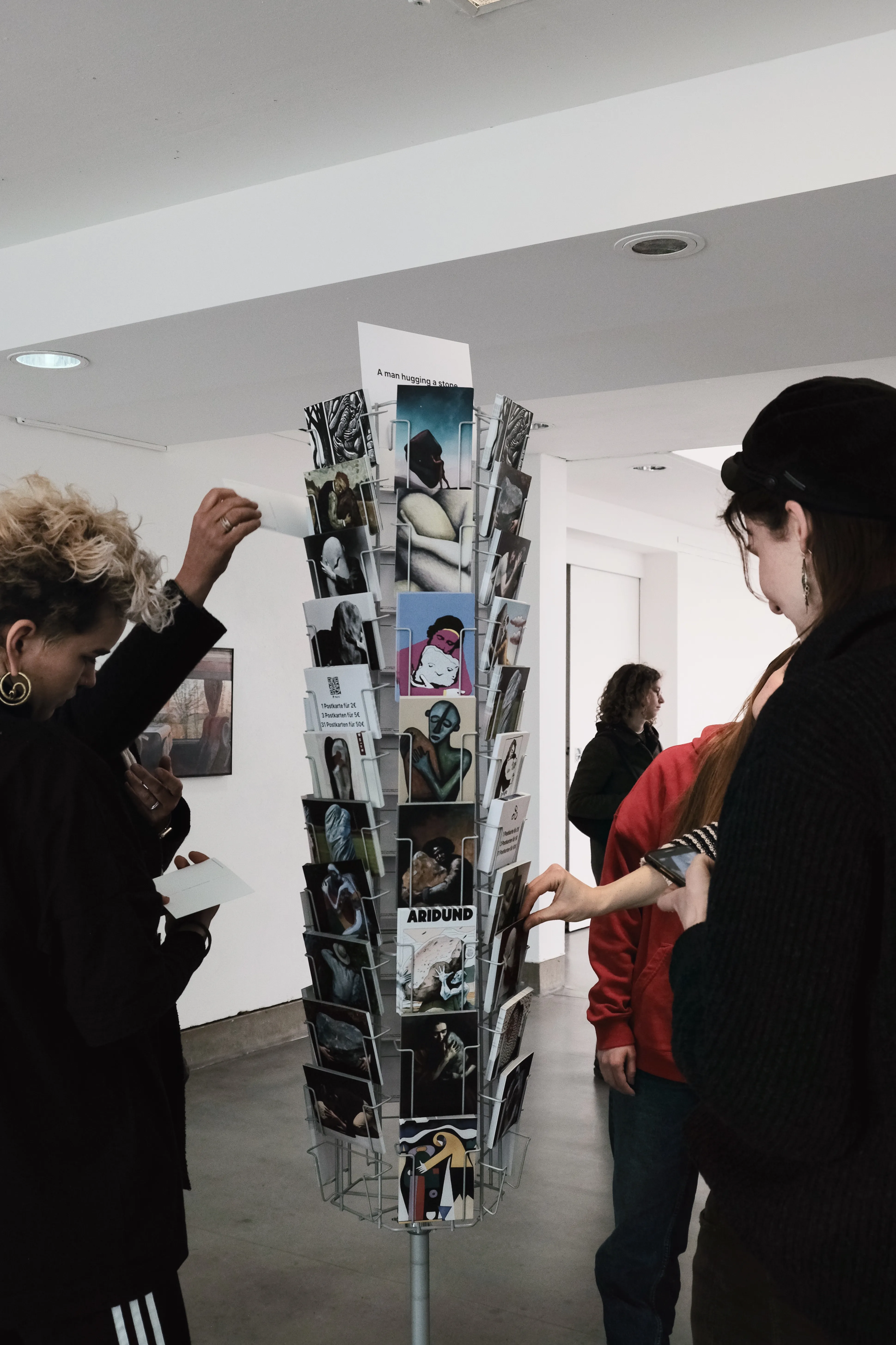 People interacting with a rotating postcard stand in an art exhibition. The stand holds various illustrated and photographic postcards, including images of figures embracing stones. A sign at the top reads 'A man hugging a stone.'