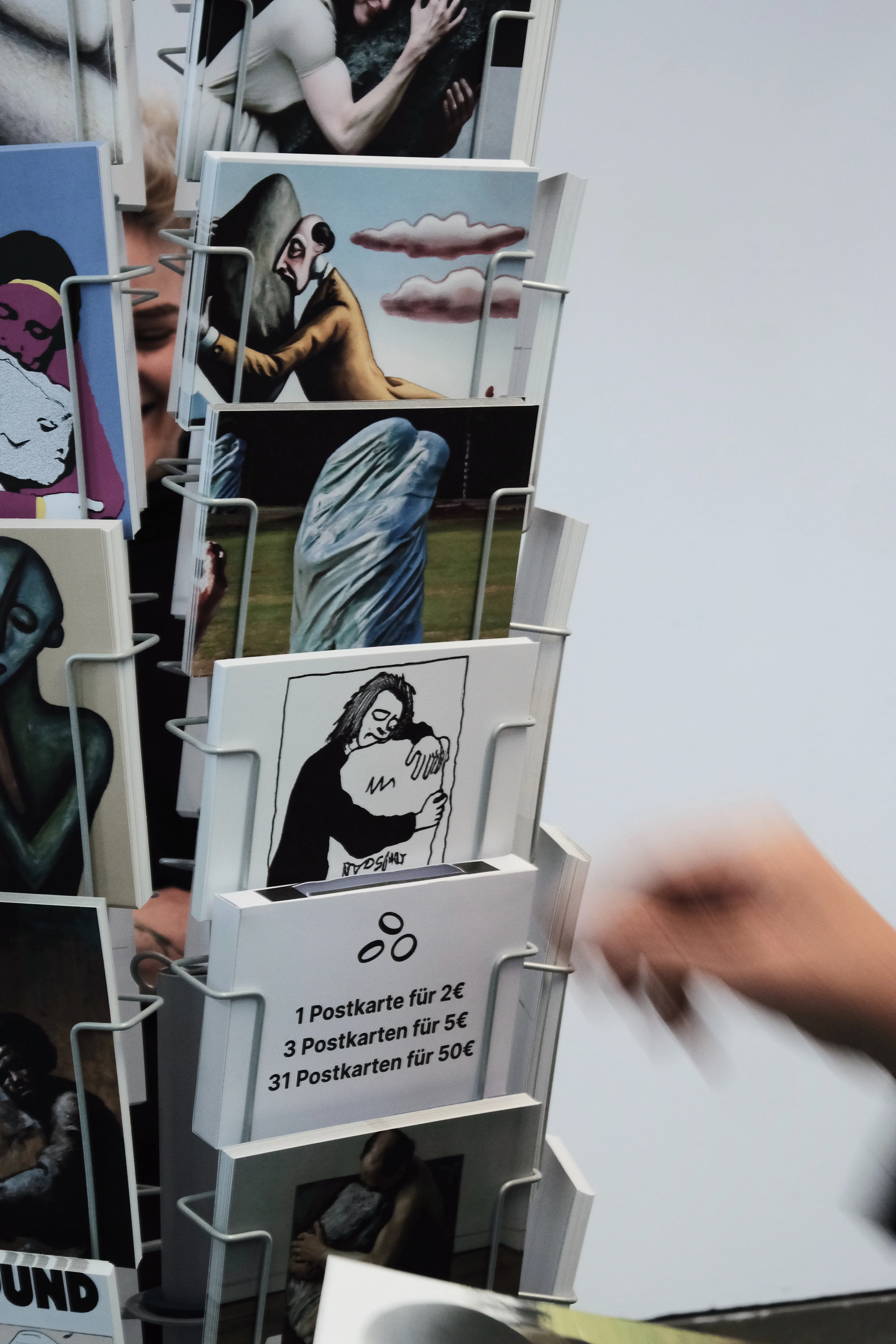 Close-up of a postcard stand displaying various artworks, including surreal and abstract depictions of figures embracing stones. A thin cardboard box the size of a postcard with an opening at the top is placed in the stand and shows an illustration of three coins and lists prices: 1 postcard for 2€, 3 for 5€, and 31 for 50€. A blurred hand is reaching for the box.