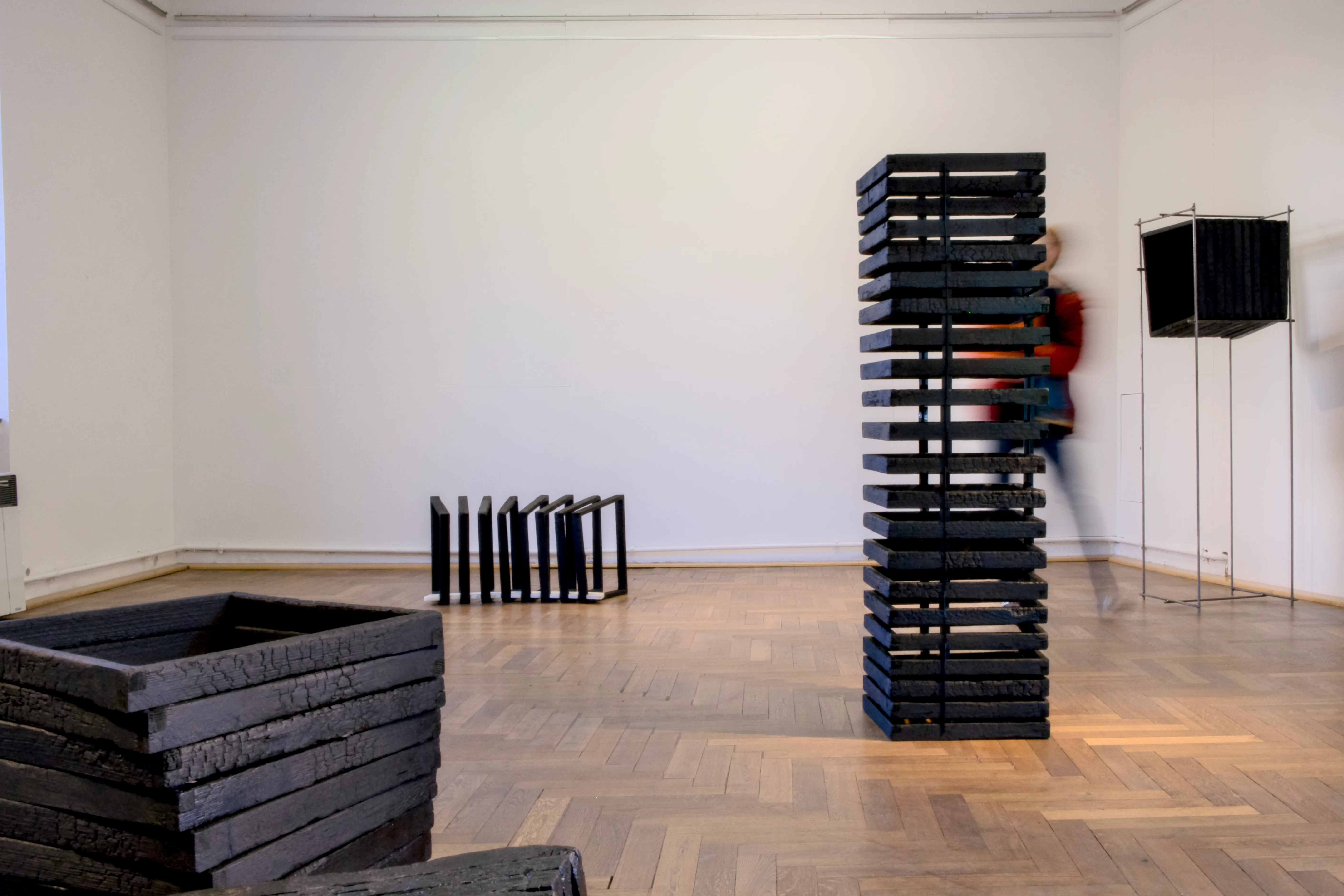 An art installation in a gallery space featuring blackened wooden squares arranged in different formations. A tall, stacked tower of squares stands in the center, while other pieces are placed on the floor and within a metal frame. A blurred figure moves behind the tower, adding a sense of motion.