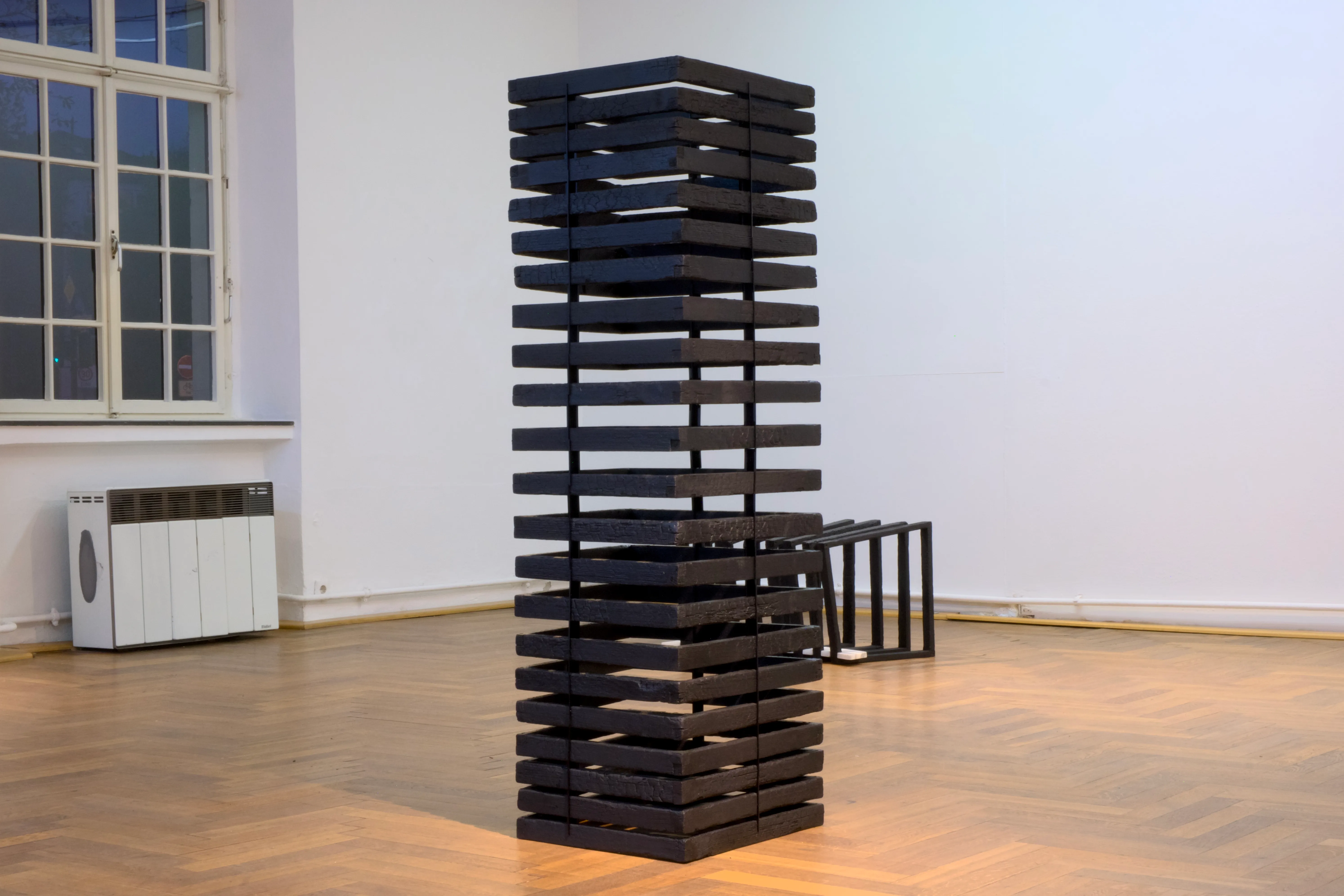 Tall vertical sculpture made of stacked, burned wooden beams forming a grid-like tower gapped by black acrylic panes in a gallery space.