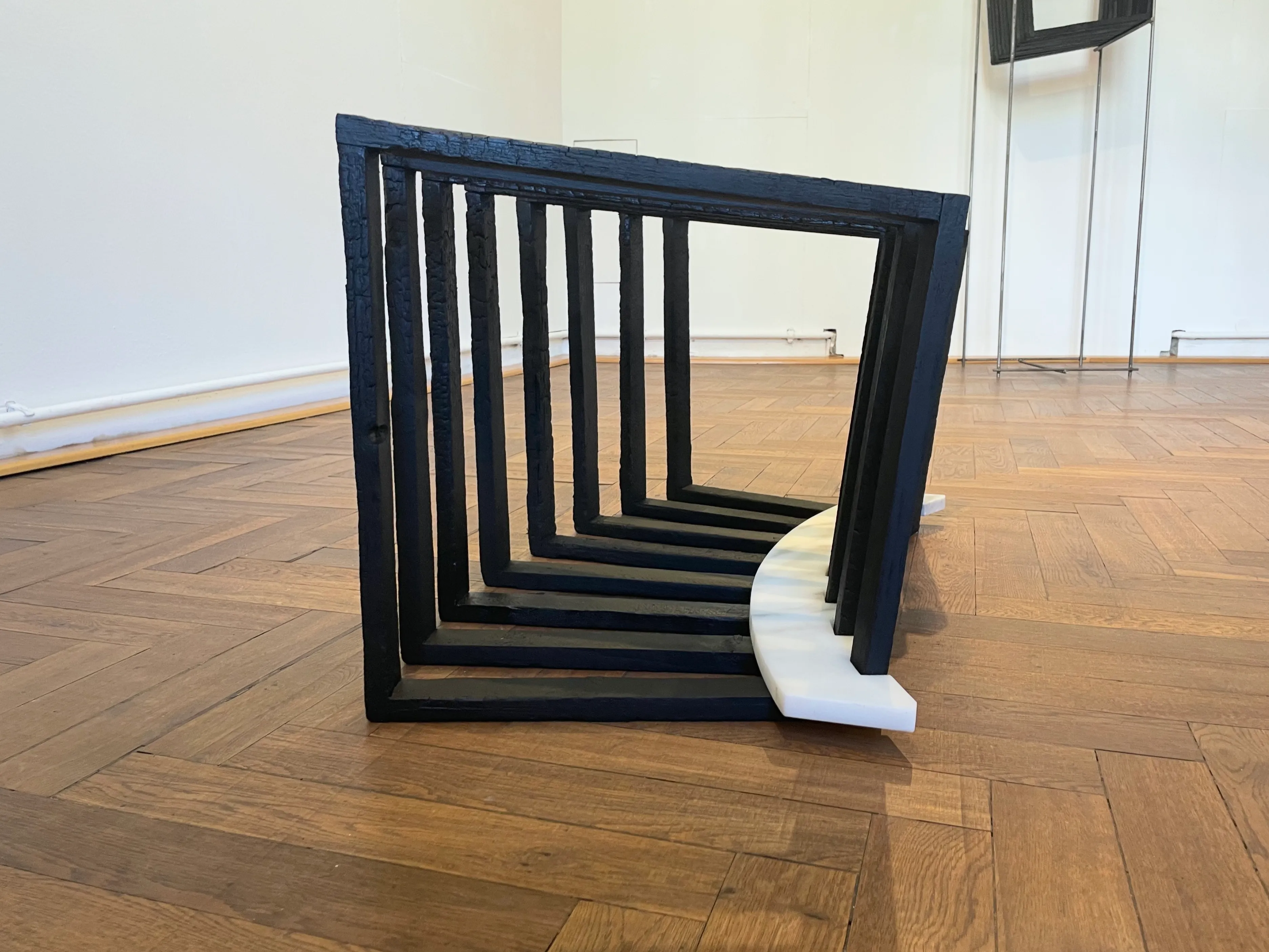 Perspective view of nested, charred wooden frames arranged in a semicircular formation on a white marble base.