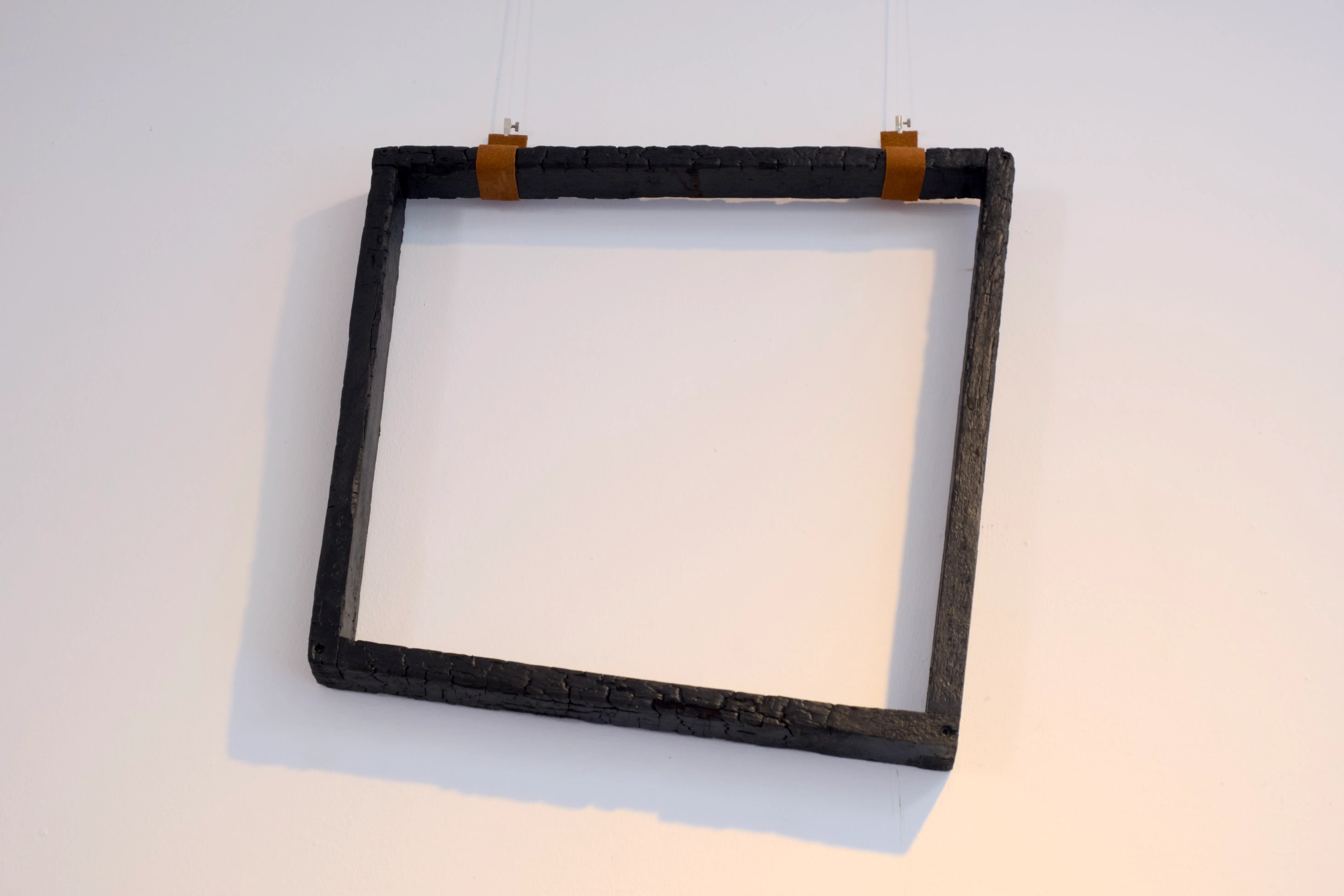 Charred wooden square frame suspended on a white wall using leather straps and thin transparent cords.
