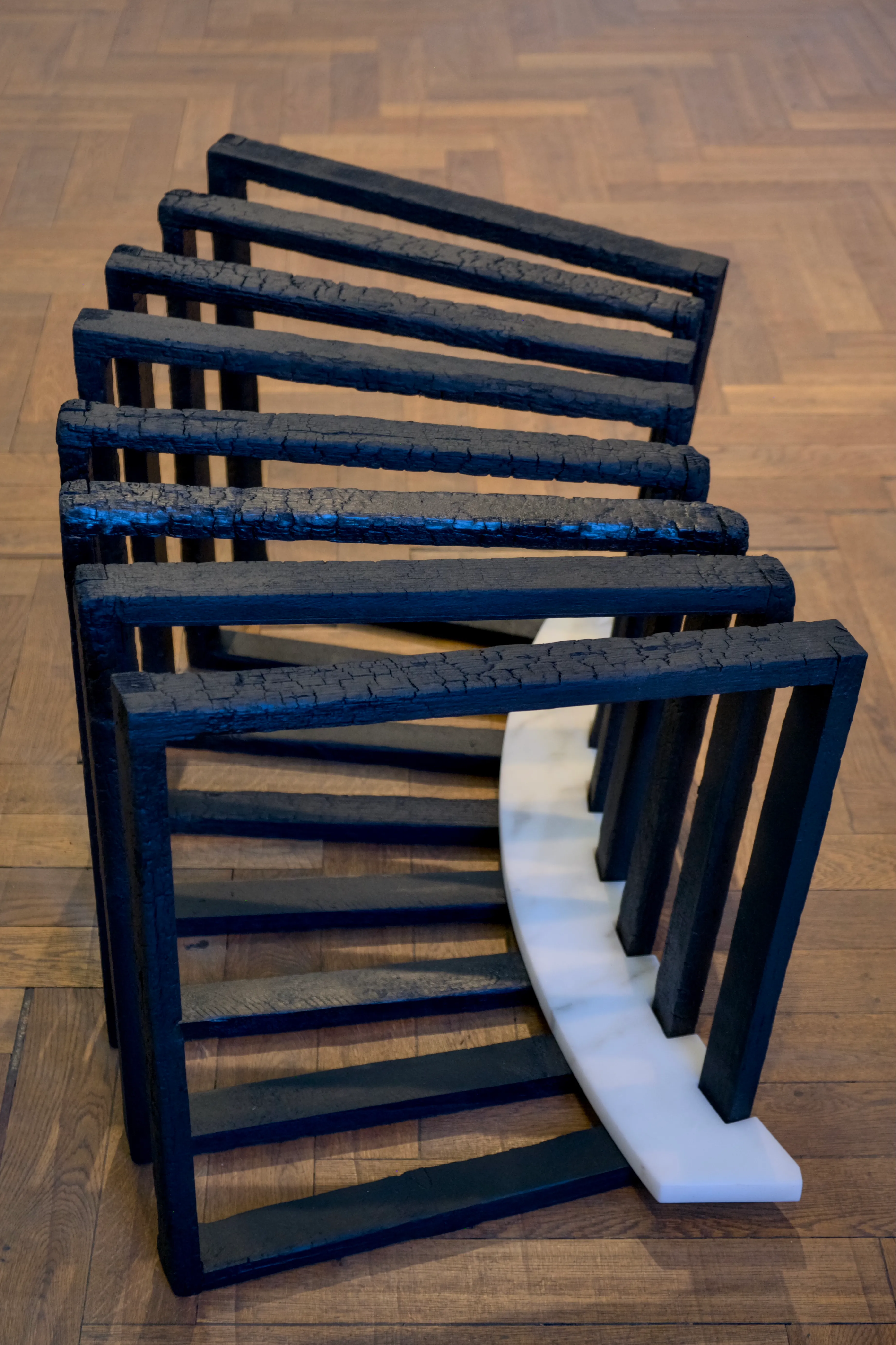 A close-up of a sculpture featuring a sequence of blackened wooden frames arranged in a curved formation. The charred surfaces display cracked textures, and the frames are supported by a white marble base, contrasting with the dark wood. The sculpure is set on a wooden parquet floor.