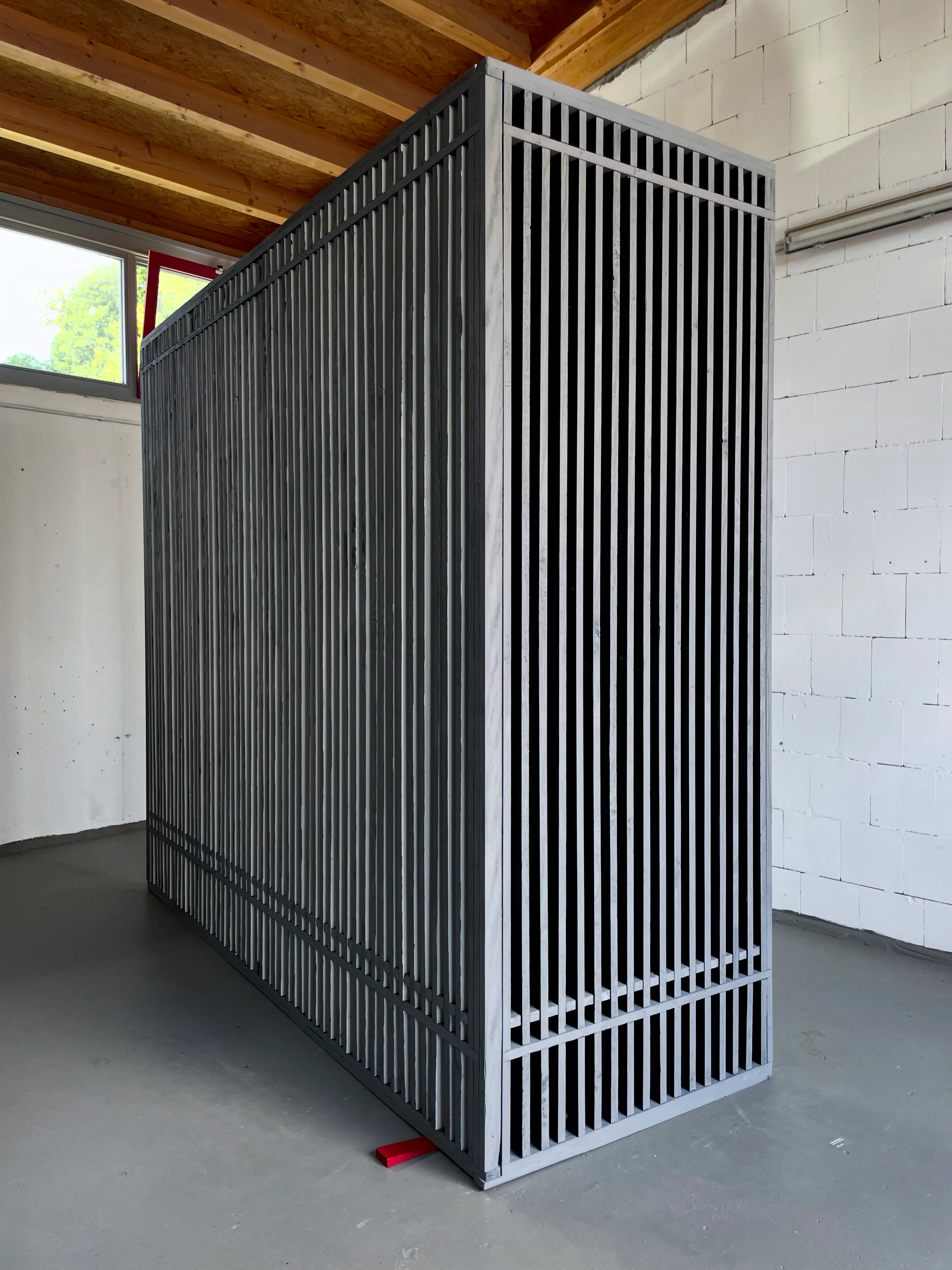 Angled view of a large rectangular sculptural wall in an L-shape made of vertical wooden slats, painted grey on the front, white on one side and black on the ither. The interleaved structure reveals glimpses of the interior through the narrow gaps. The longer part is prominently supported neear the corner by a red wooden wedge.