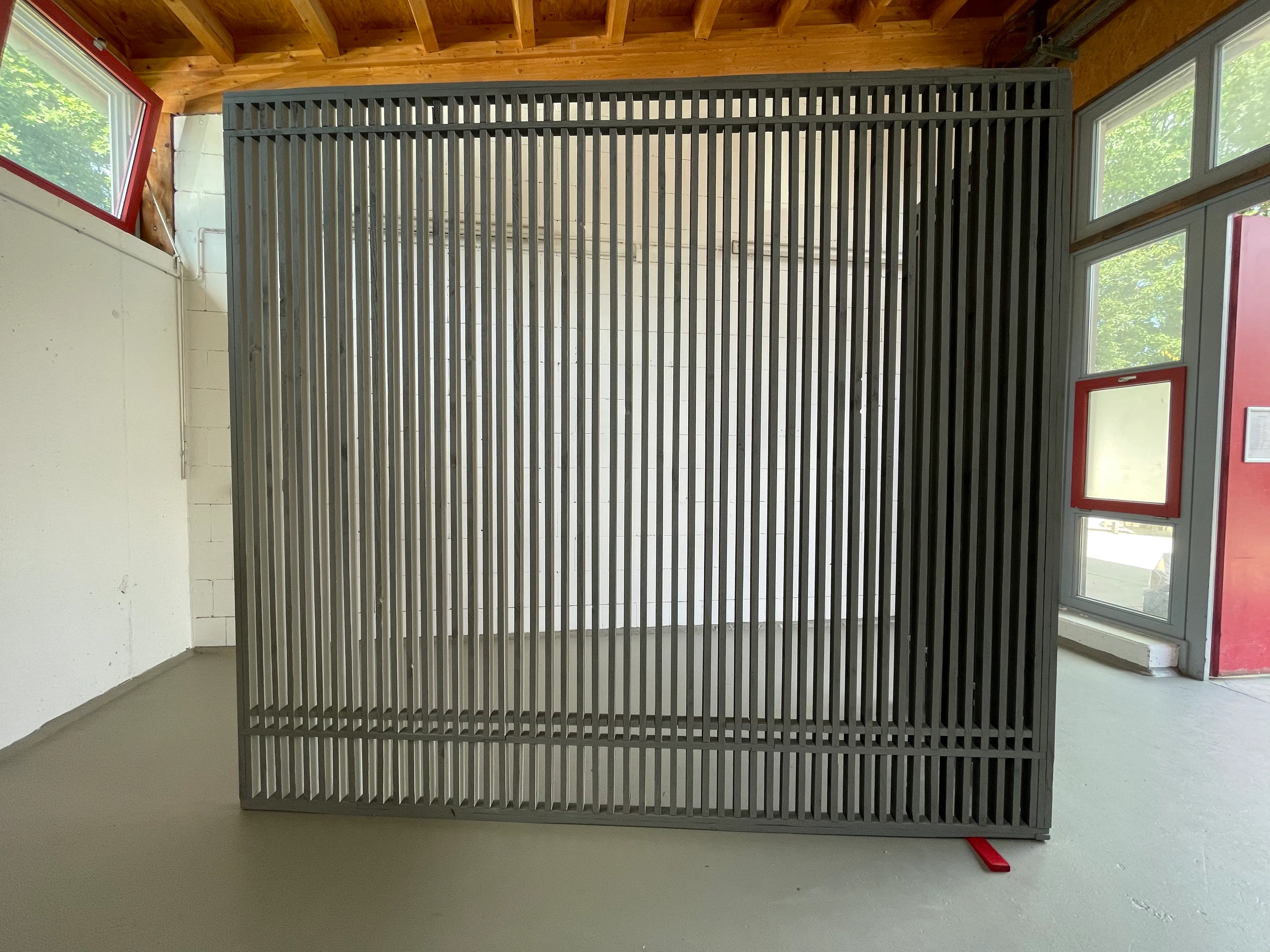 Side view of the installation with evenly spaced wooden slats creating a striped visual effect. The black and white sides of the slats produce a dynamic, optical contrast. The longer part is prominently supported near the corner by a red wooden wedge.