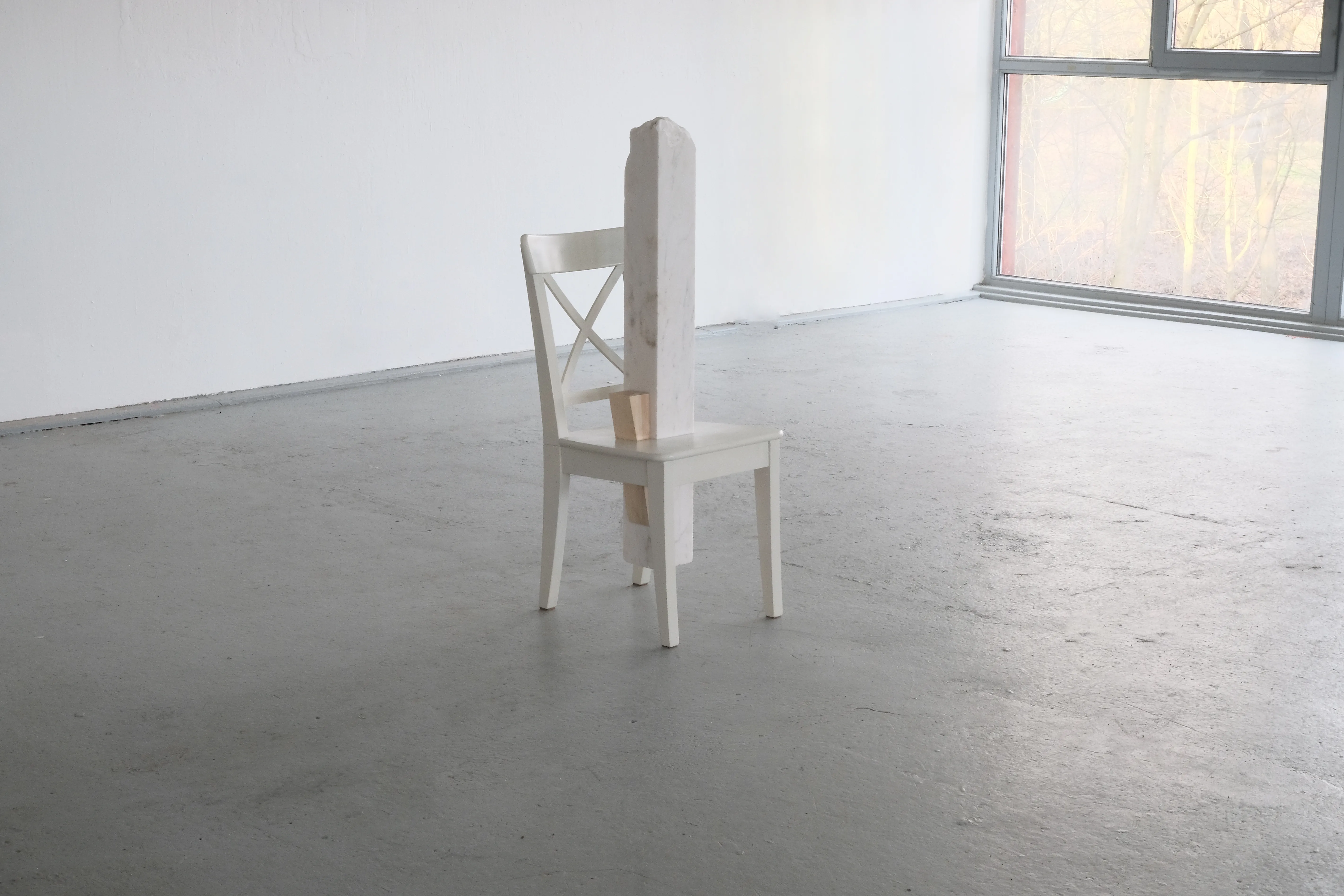 A white wooden chair stands alone in a minimalistic gallery space with a large window in the background. A vertical block of white marble is inserted through the seat and extends upwards, partially obstructing the chair’s backrest. A smaller wooden wedge supports the marble block, creating a contrast between the materials.