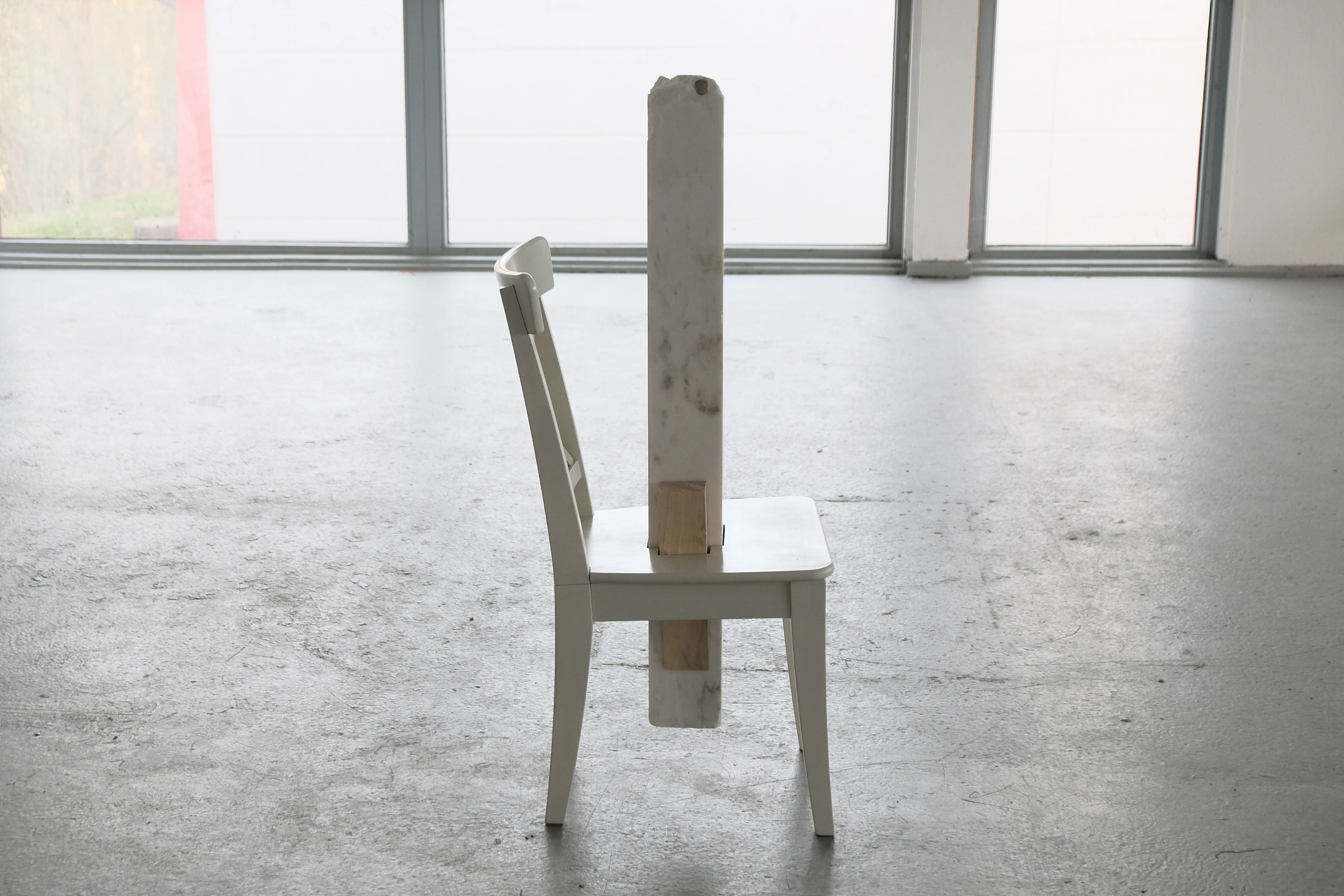 A white wooden chair stands alone in a minimalistic gallery space with a large window in the background. A vertical block of white marble is inserted through the seat and extends upwards, partially obstructing the chair’s backrest. A smaller wooden wedge supports the marble block, creating a contrast between the materials.