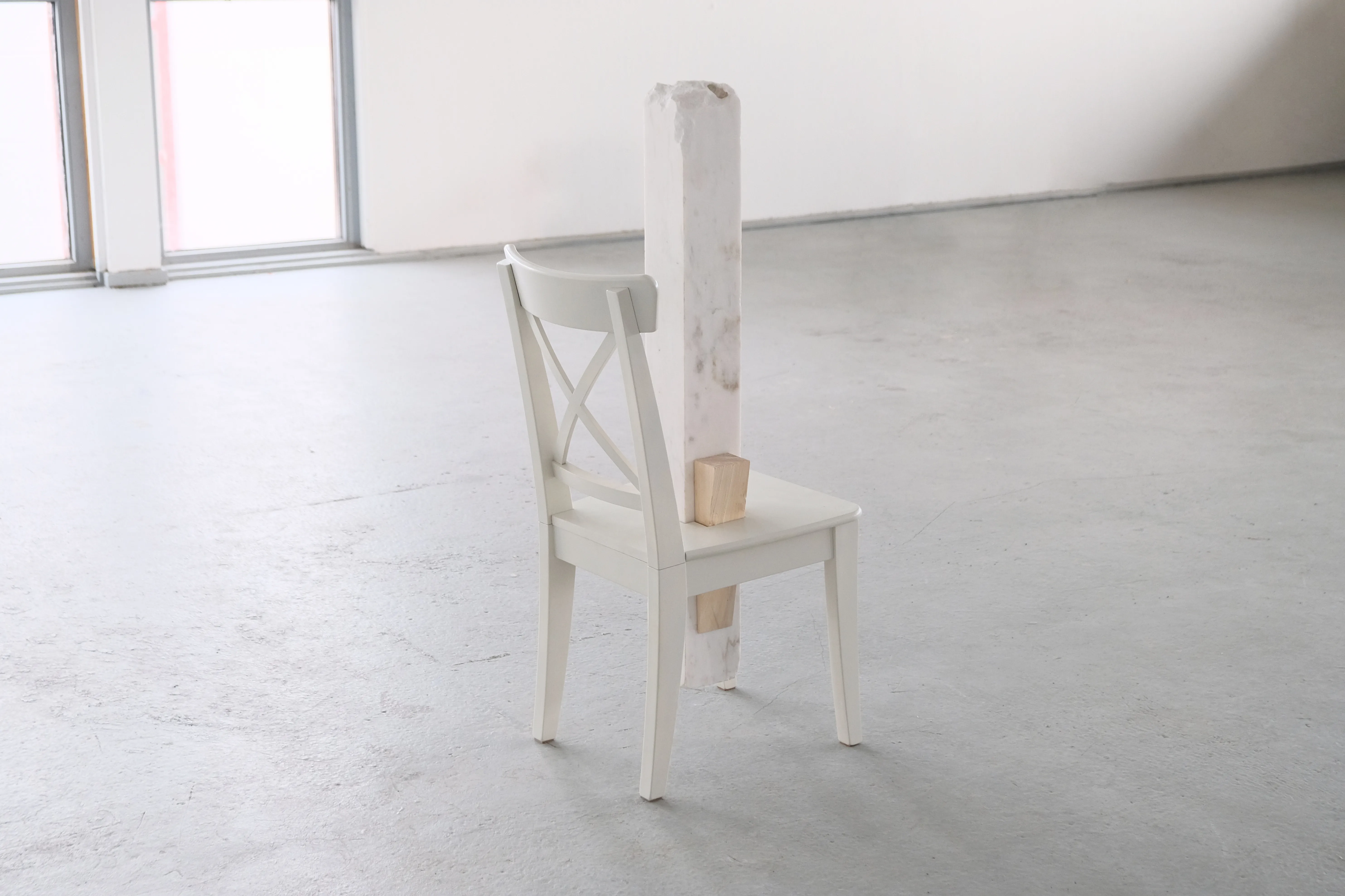 A white wooden chair stands alone in a minimalistic gallery space with a large window in the background. A vertical block of white marble is inserted through the seat and extends upwards, partially obstructing the chair’s backrest. A smaller wooden wedge supports the marble block, creating a contrast between the materials.