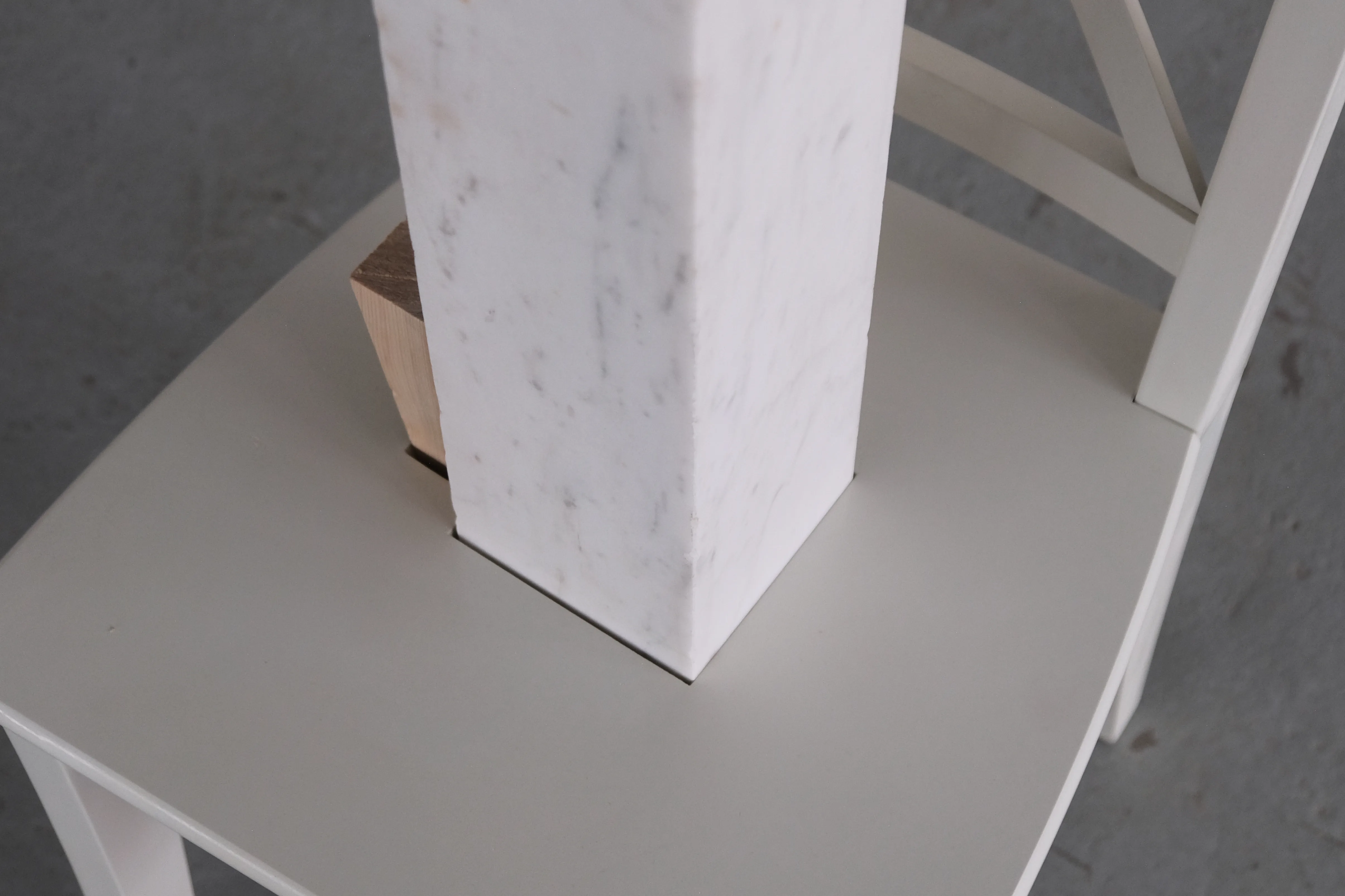 Close-up of a white chair sculpture with a square hole in the seat, through which a white marble block extends. A small wooden wedge is placed at the base of the block, contrasting the smooth surfaces of the chair and marble.