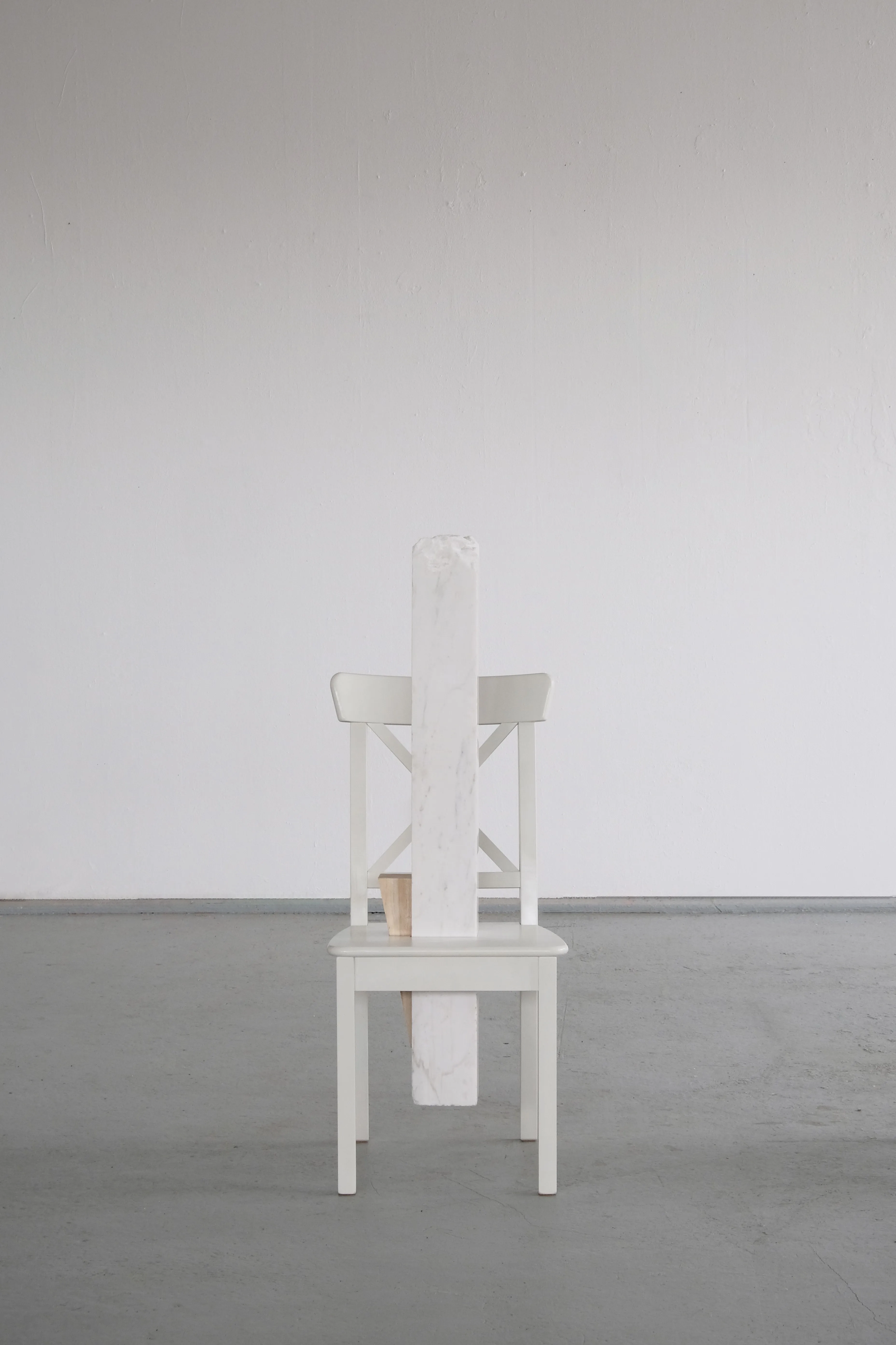 A white wooden chair stands alone in front of a white wall. A vertical block of white marble is inserted through the seat and extends upwards, partially obstructing the chair’s backrest. A smaller wooden wedge supports the marble block, creating a contrast between the materials.